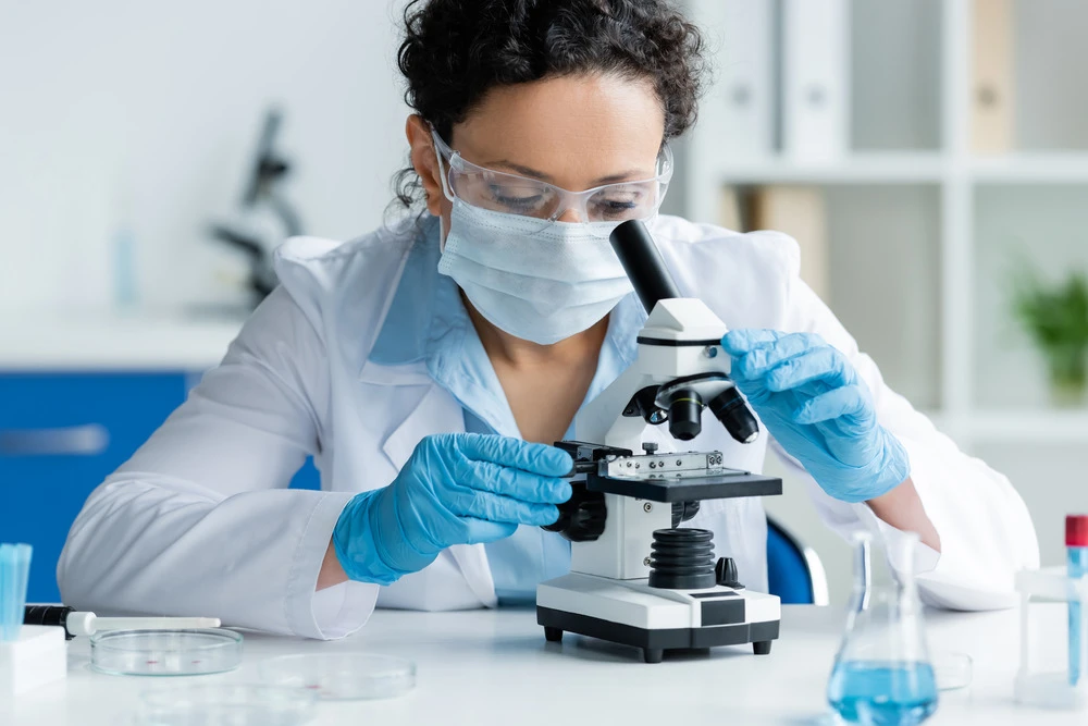 African american scientist in medical mask and latex gloves looking through microscope near petri dishes