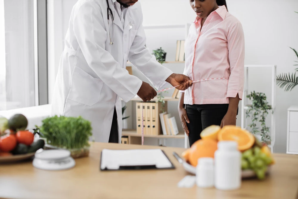 Cropped view of multicultural man in lab coat holding measuring tape on woman's waist in doctor's office.