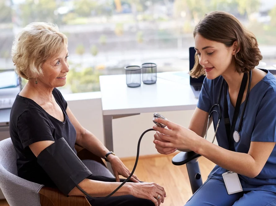 Nurse Wearing Scrubs In Office Checking Senior Female Patients Blood Pressure