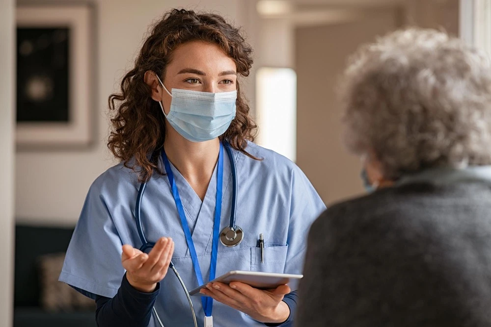Doctor wearing safety protective mask supporting and cheering up senior patient during home visit