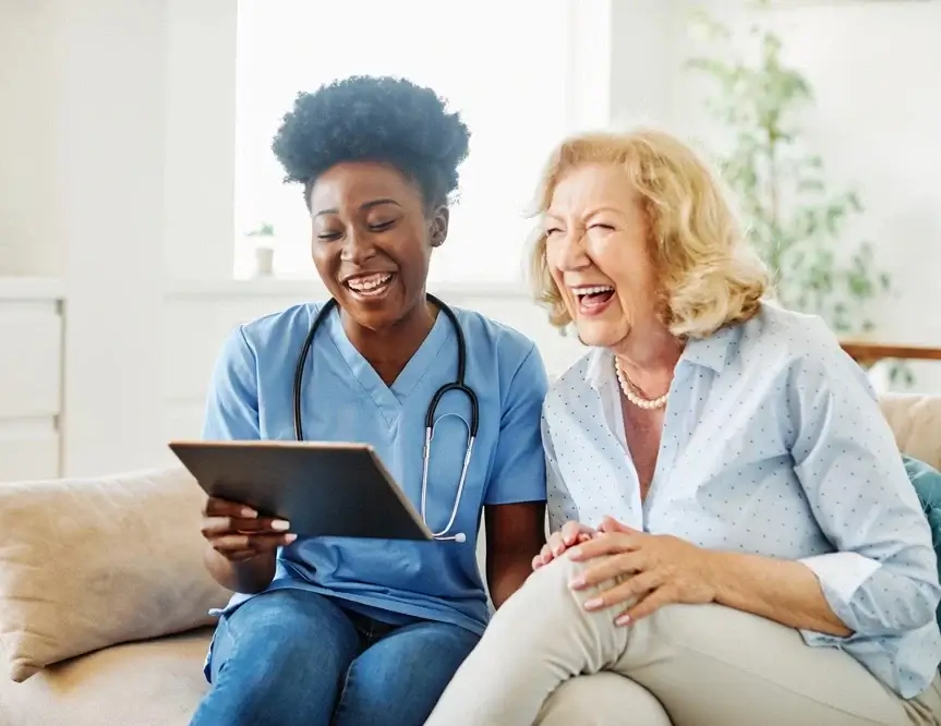 Doctor or nurse caregiver showing a tablet screen to senior woman and laughing at home