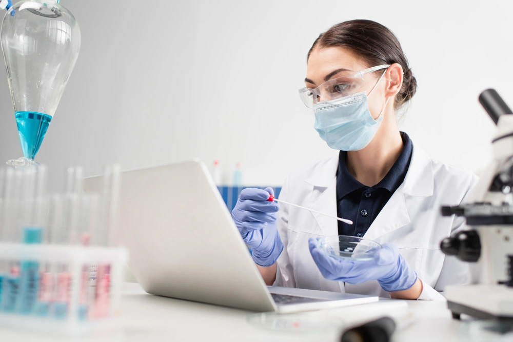 Scientist in medical mask holding pcr test and petri dish near laptop and microscope in lab