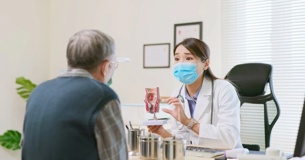 asian female doctor wearing face mask is showing a rectal model and explaining to elder senior man patient in hospital