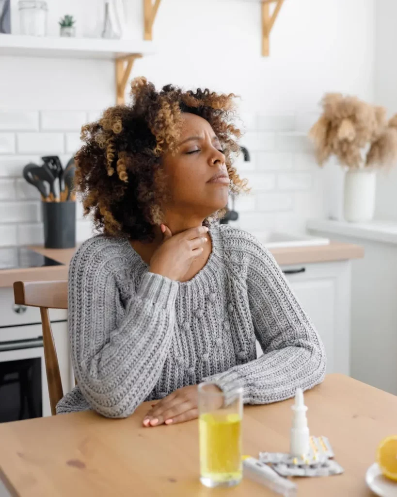 African American woman with a cold and sore throat drinks vitamins and measures body temperature.